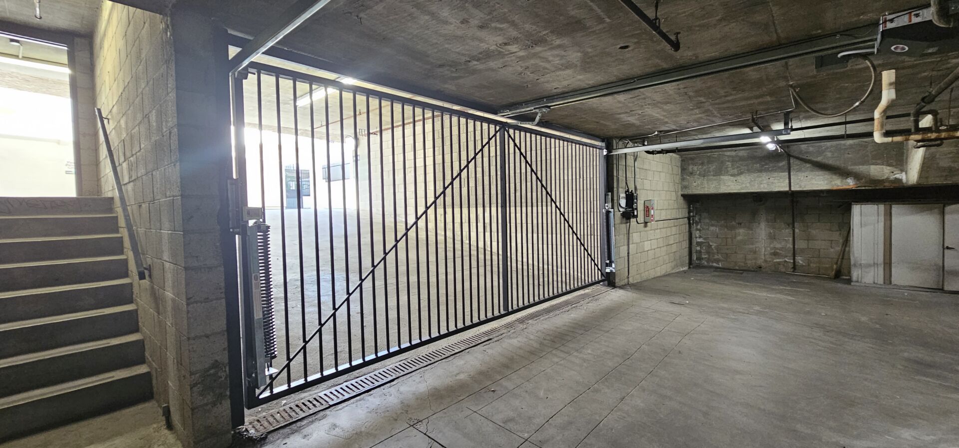 Barred steel sliding security gate on a floor track inside an underground parking garage, with a stairwell alongside