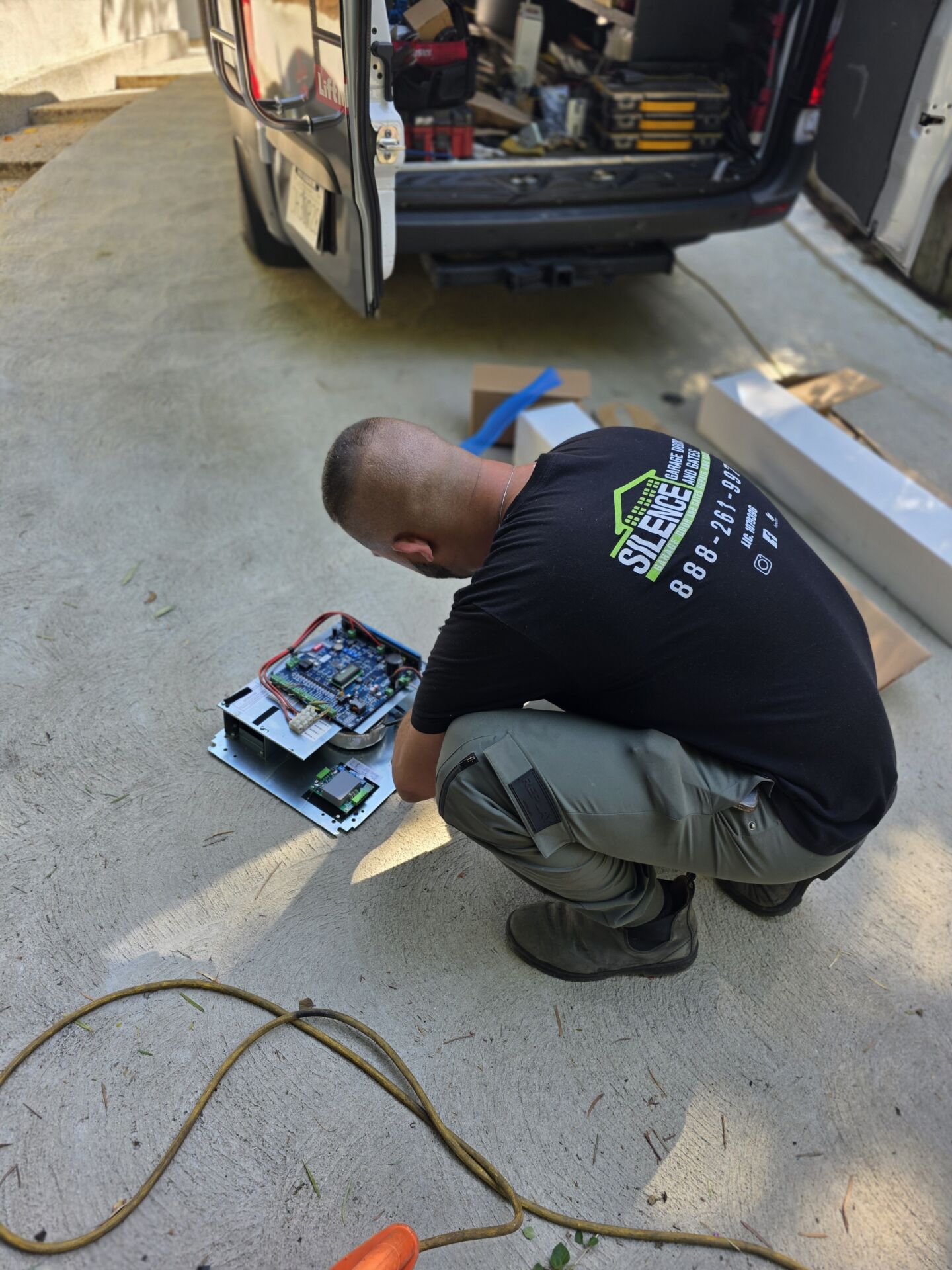 Technician kneeling on the ground working on a gate control board laid out beside an open service van