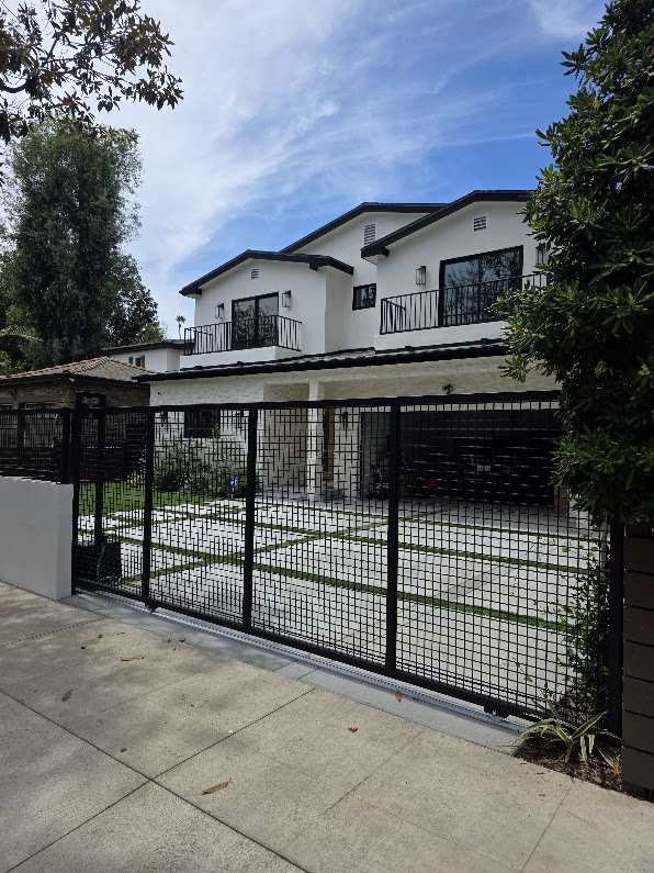 Black steel mesh sliding gate closed across a ribbon driveway on a bottom track, with a two-storey white house behind