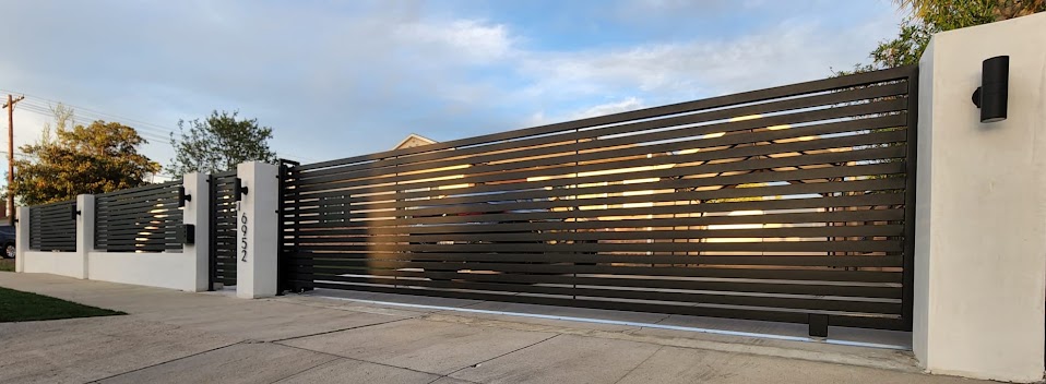 Wide view of a completed dark horizontal slat sliding driveway gate between white stucco columns, with a matching pedestrian gate alongside