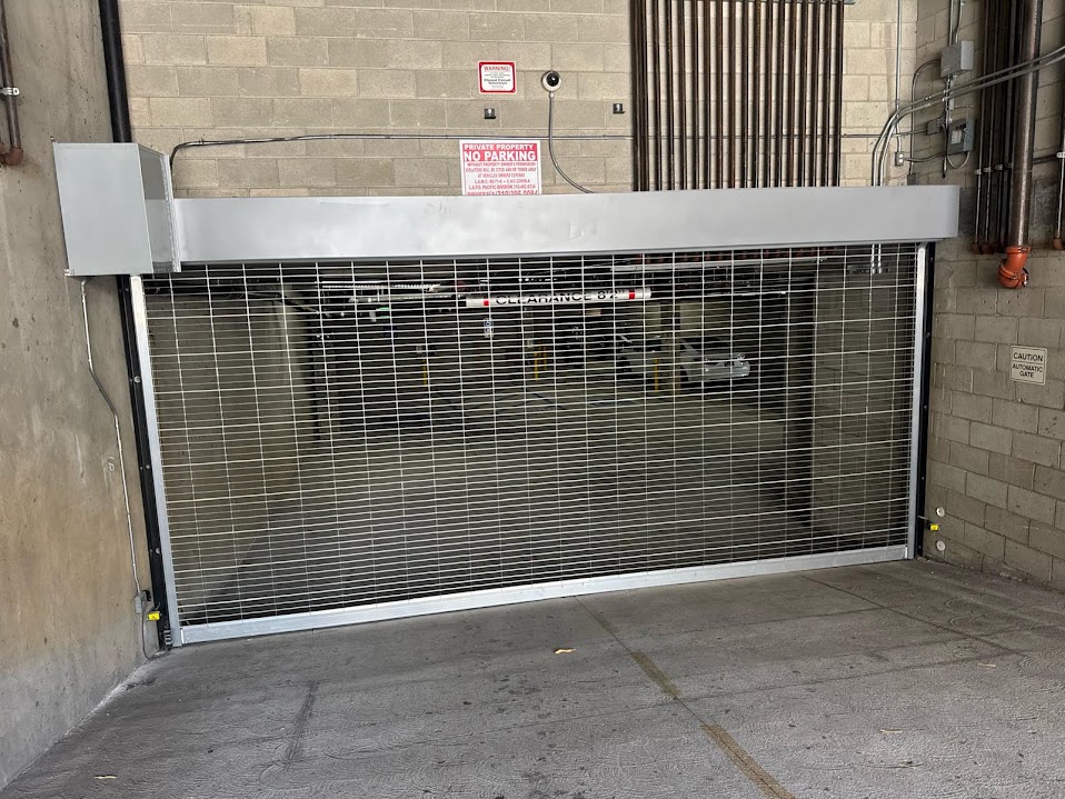 Galvanised steel mesh rolling security grille partly closed across an underground parking garage entrance, with its coil housing above