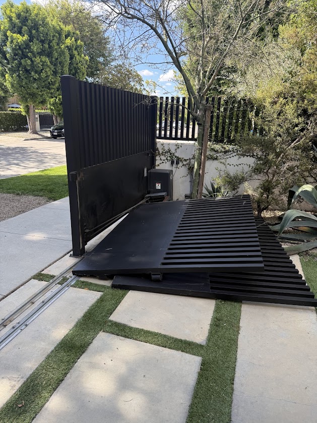 A sliding driveway gate off its track, lying on the paver walkway beside the opening