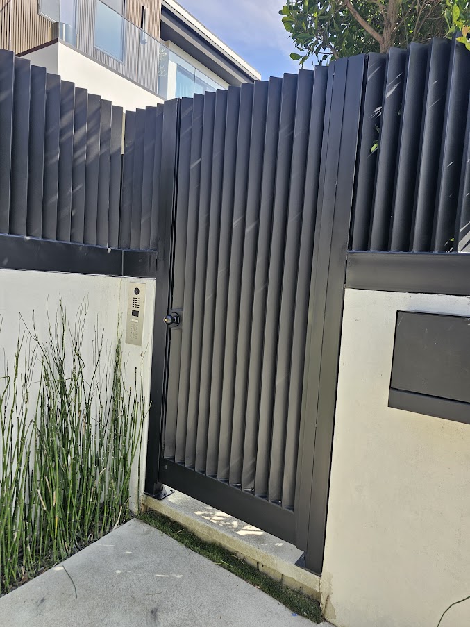 Vertical-slat metal pedestrian gate with DoorBird intercom, set in a smooth stacker-block wall. West Hollywood.