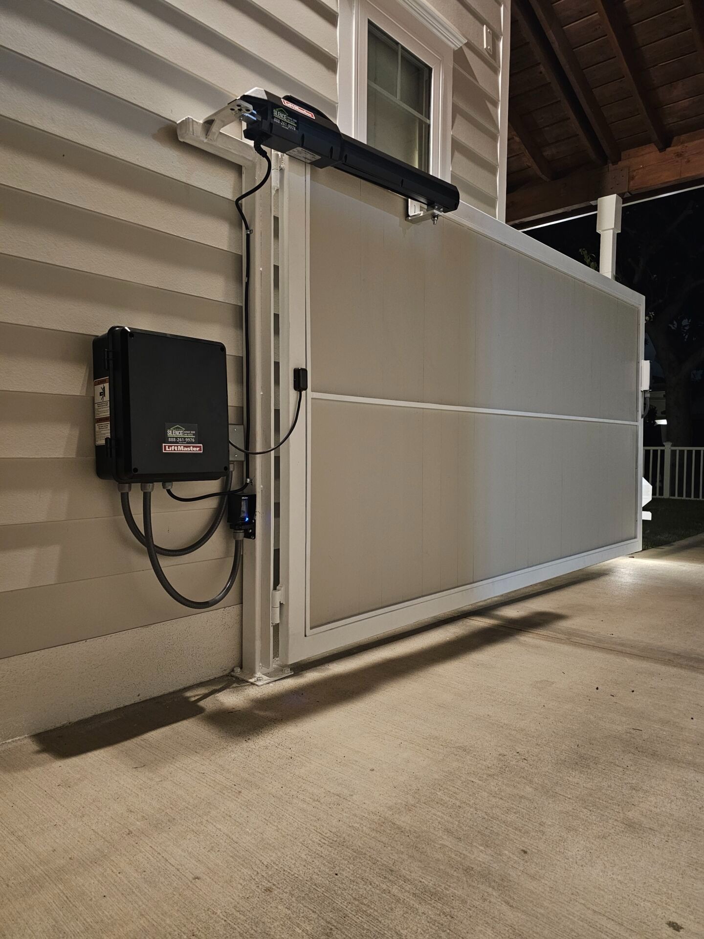 LiftMaster swing gate operator and control box mounted beside a gate leaf, with a photo-eye safety sensor on the post