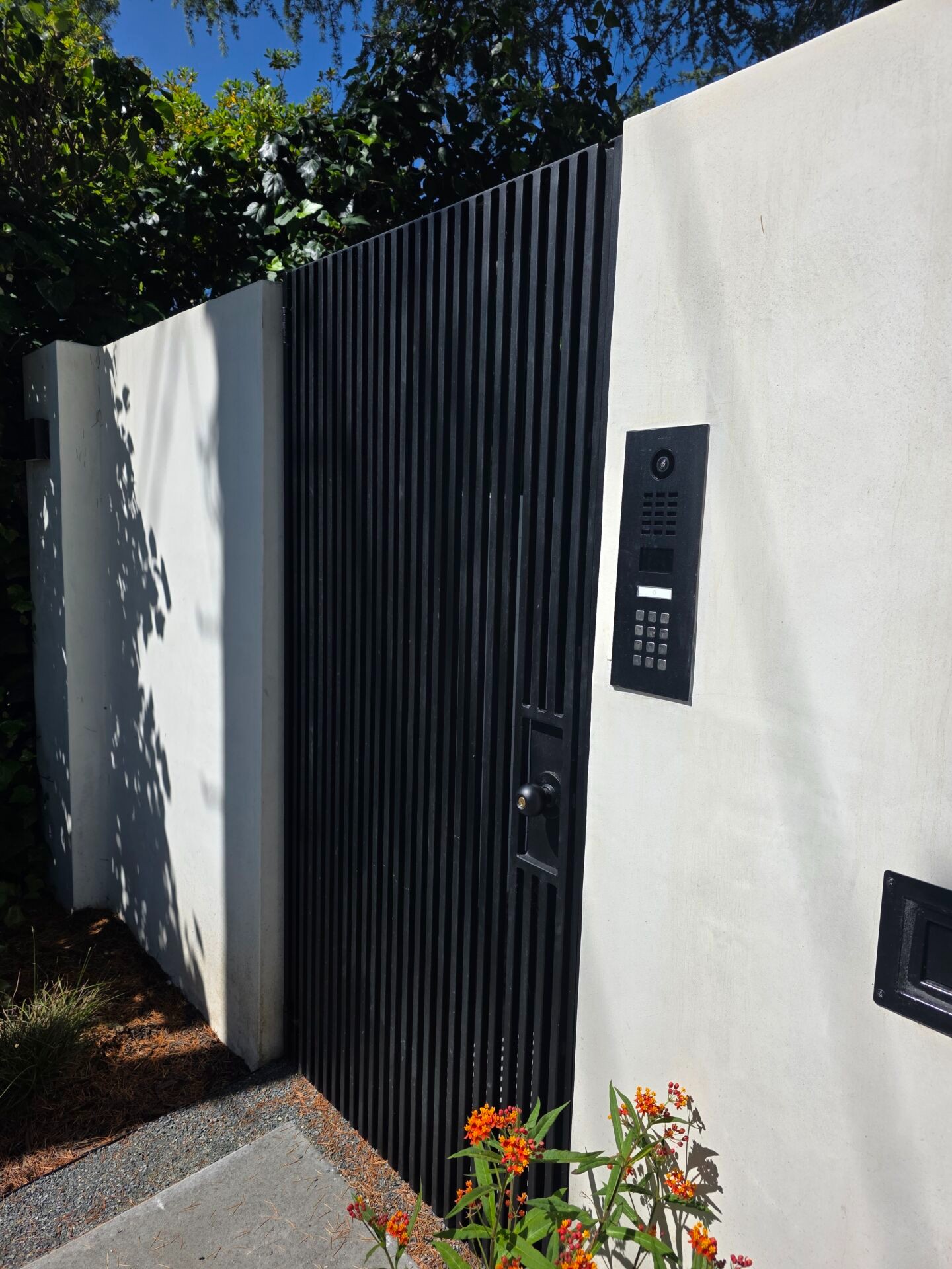 DoorBird video intercom with keypad mounted on a stucco wall beside a black vertical-slat gate