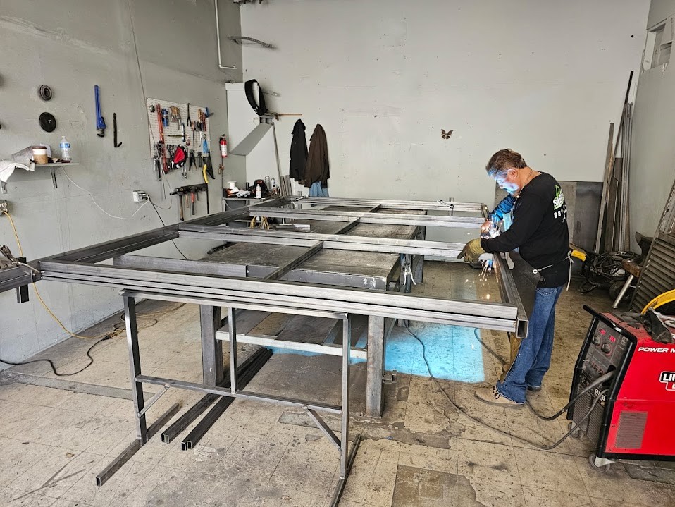 Steel gate frame laid out on a welding table in the fabrication shop while a fabricator welds a joint
