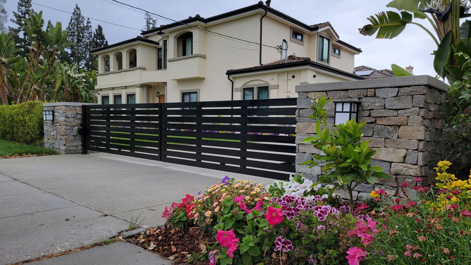 New black horizontal-slat sliding driveway gate and matching fence set into a stacked-stone wall in front of a two-story home