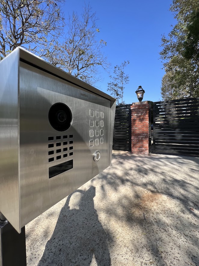 Stainless steel DoorBird video intercom with keypad mounted on a post beside a slat gate and brick column