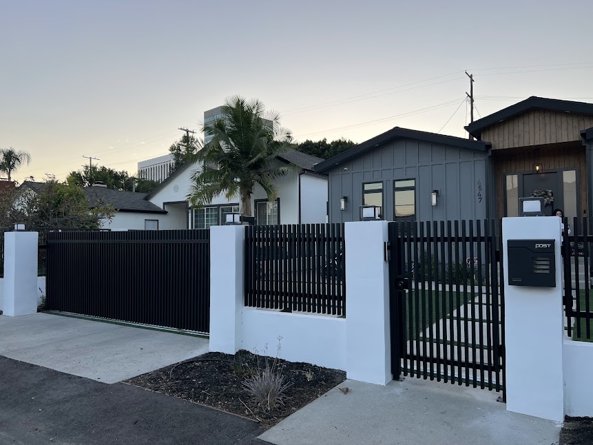 New black vertical-slat sliding driveway gate with a matching pedestrian gate, fence and white smooth-stack columns
