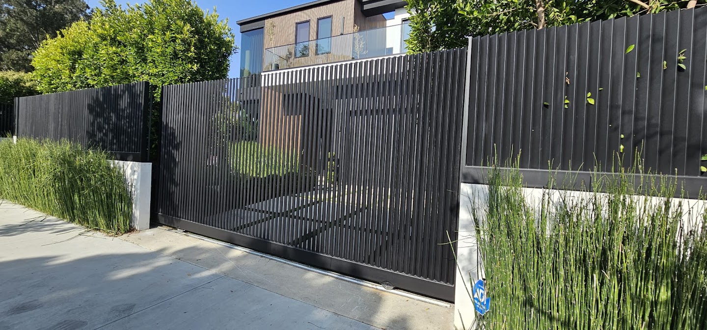 New dark vertical-slat sliding driveway gate and matching fence above a smooth block wall with planting alongside